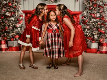 Little girl gets kisses from sisters in red, in front of Christmas trees and red fireplace backdrop
