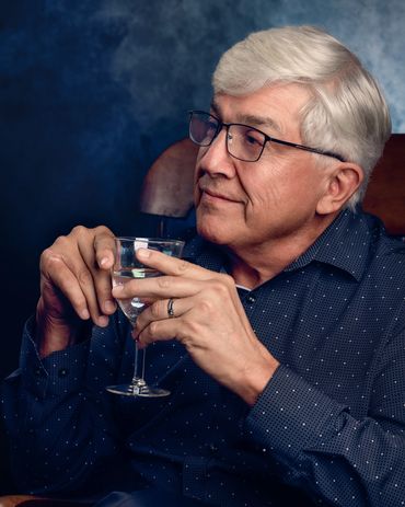 Older man with white hair, glasses, blue shirt holds wine glass in front of blue backdrop