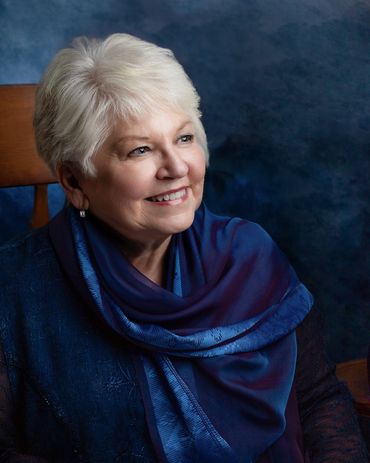 Lovely older woman with white hair, draped in blue, sits in rocking chair in front of blue backdrop