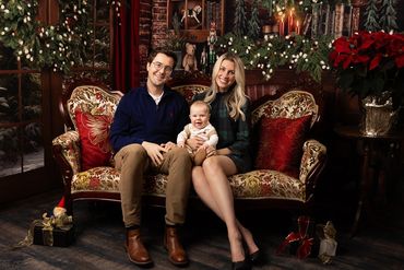 Young parents sit in decorated Christmas room on Victorian sofa with their infant baby.