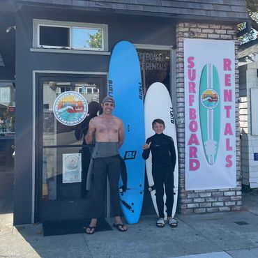 Two people with surfboards outside a surfboard rental shop.
