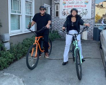 Two people smiling on e-bikes in front of a rental shop.
