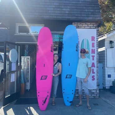 Two people holding colorful surfboards outside a rental shop on a sunny day.