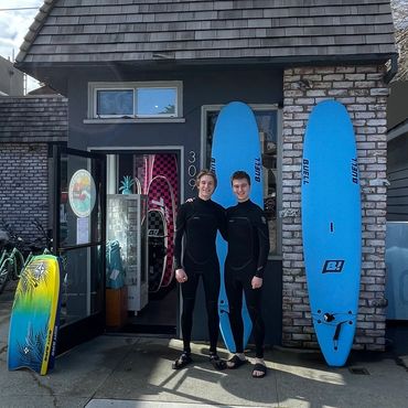 Two surfers in wetsuits stand outside a surf shop with blue surfboards.