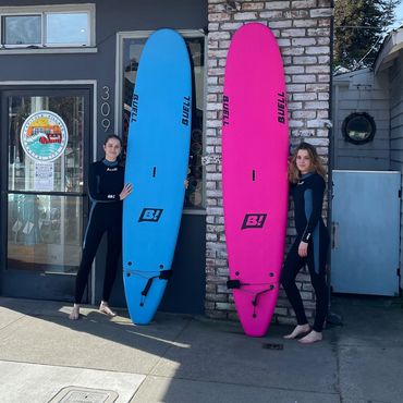 Two surfers in wetsuits holding bright blue and pink surfboards outside a rental shop.