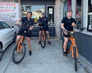 Three men posing with orange rental bikes outside a bike rental shop.