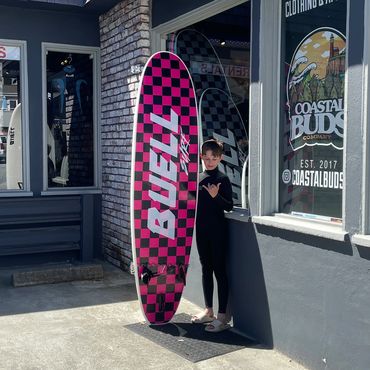 A boy in a wetsuit stands with a pink checkered Buzzz surfboard outside a coastal shop.