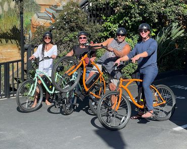 Four adults wearing helmets on bicycles, two doing wheelies, posing outdoors in bright sunlight.
