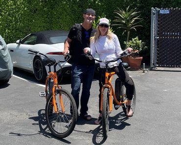 A man and woman stand with orange bicycles in a sunny parking lot.