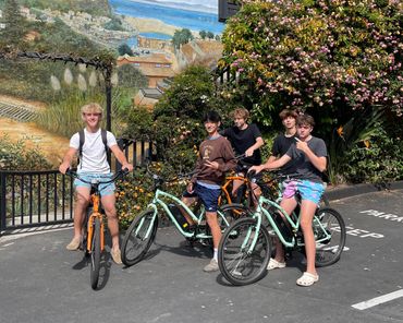 A group of five young men posing with bicycles near a mural and flowering bushes.
