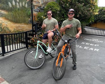 Two men posing with bicycles in front of a gate and mural.