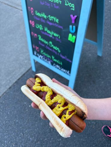 Hand holding a hot dog with mustard and ketchup in front of a menu board.