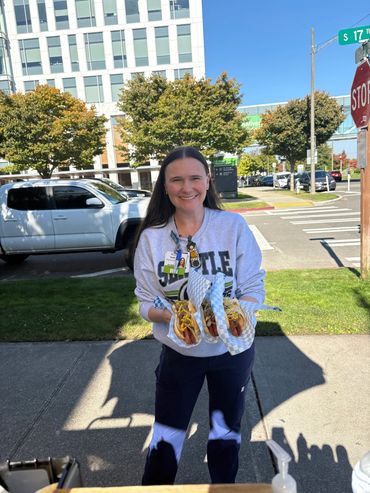 A smiling woman holds three hot dogs on a sunny street corner.
