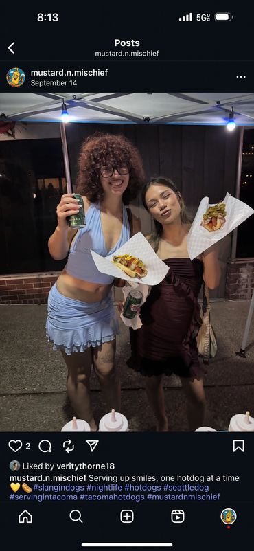 Two women smiling and holding hotdogs and drinks at night under a tent.