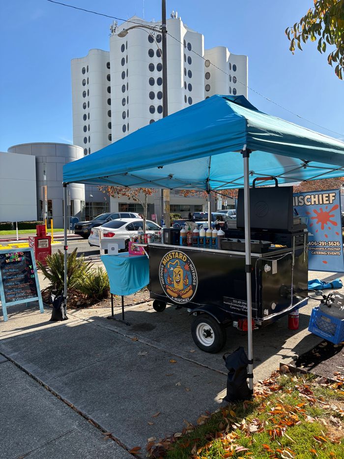 Hotdog stand with blue canopy set against a modern building on a sunny day.