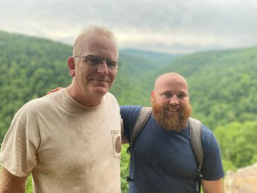 Two men smiling with forested hills in the background.