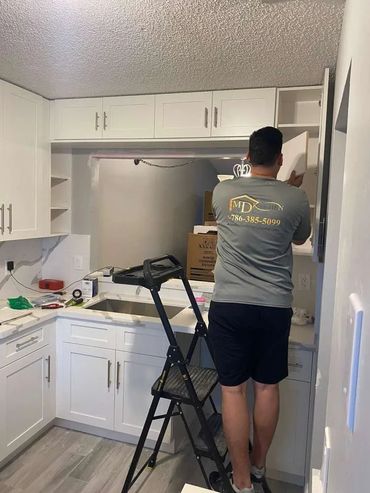 A man installs or inspects white kitchen cabinets using a step ladder.