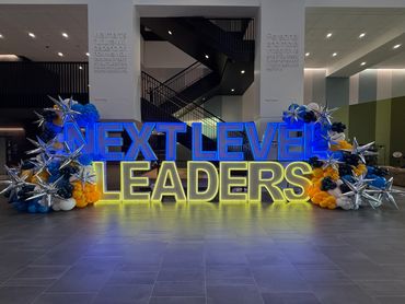 Illuminated "Next Level Leaders" sign with decorative balloons and stars in a modern lobby.
