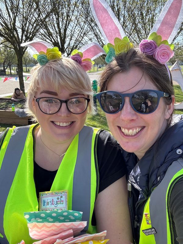 Two women wearing bunny ears and reflective vests smile outdoors on a sunny day.