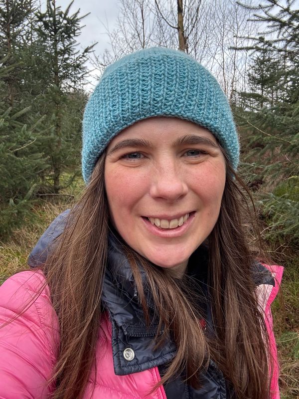 Smiling woman in a blue knit hat and pink jacket outdoors in a forest.