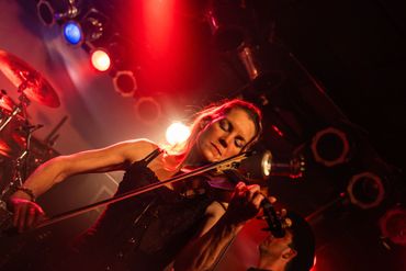 Violinist playing passionately on stage with red lighting and band members.