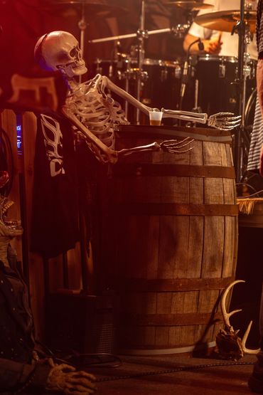 Skeleton sitting at a wooden barrel with a drink in a dimly lit music venue.