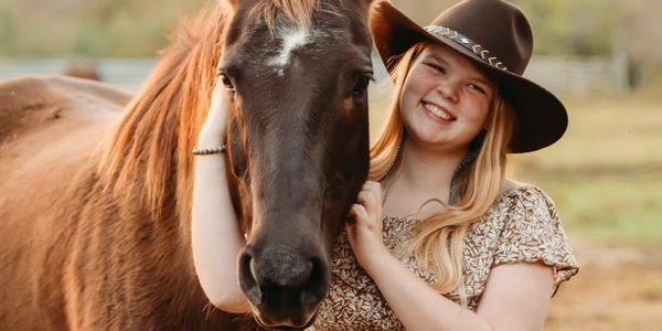A smiling young woman in a cowboy hat embraces a brown horse outdoors.