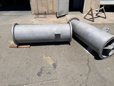 Two large industrial metal pipes that were sandblasted, lying on concrete outside a warehouse.