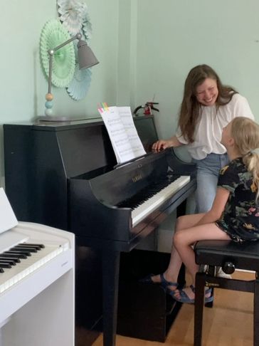 A music teacher smiling at a young student by the piano.