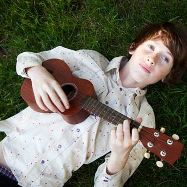 Boy lying on grass playing a small guitar.
