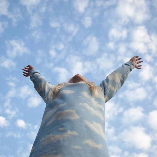 Person wearing a cloud-patterned outfit with arms raised against a cloudy sky.