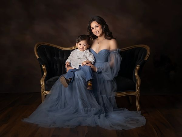 A smiling woman in a blue gown holds a toddler on a vintage sofa.