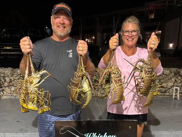Two happy people holding lobsters, posing behind a Whiskey Throttle Charters sign at night.