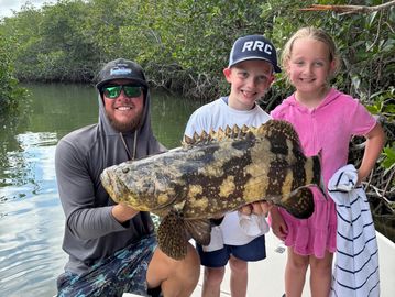 Man and two kids proudly hold a large fish on a boat by a mangrove forest.