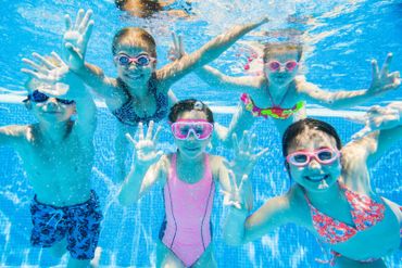 Happy kids underwater in swimming pool wearing goggles and showing hands.