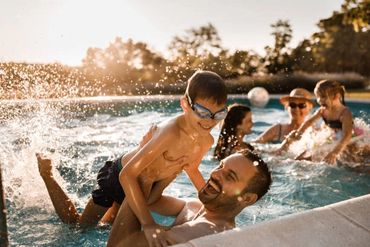 A father joyfully lifts his son in a sunlit pool with family playing in the background.