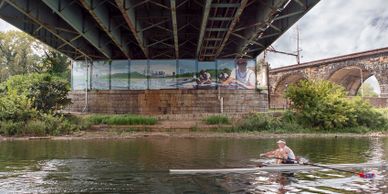 A rower on a river boats past a mural under a bridge overpass.