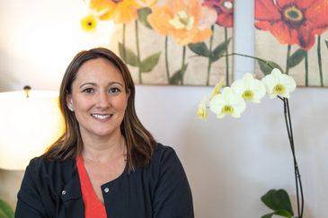 Smiling woman sitting indoors with floral artwork and orchids in the background.