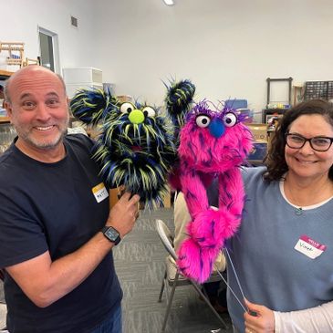 Two people holding colorful furry puppets and smiling indoors.