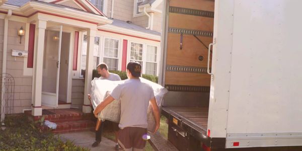 Two men carrying a mattress into a house from a moving truck.