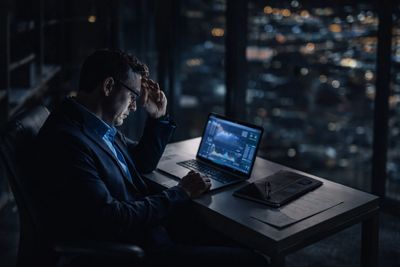 A man in a suit works late at night on a laptop with financial charts.