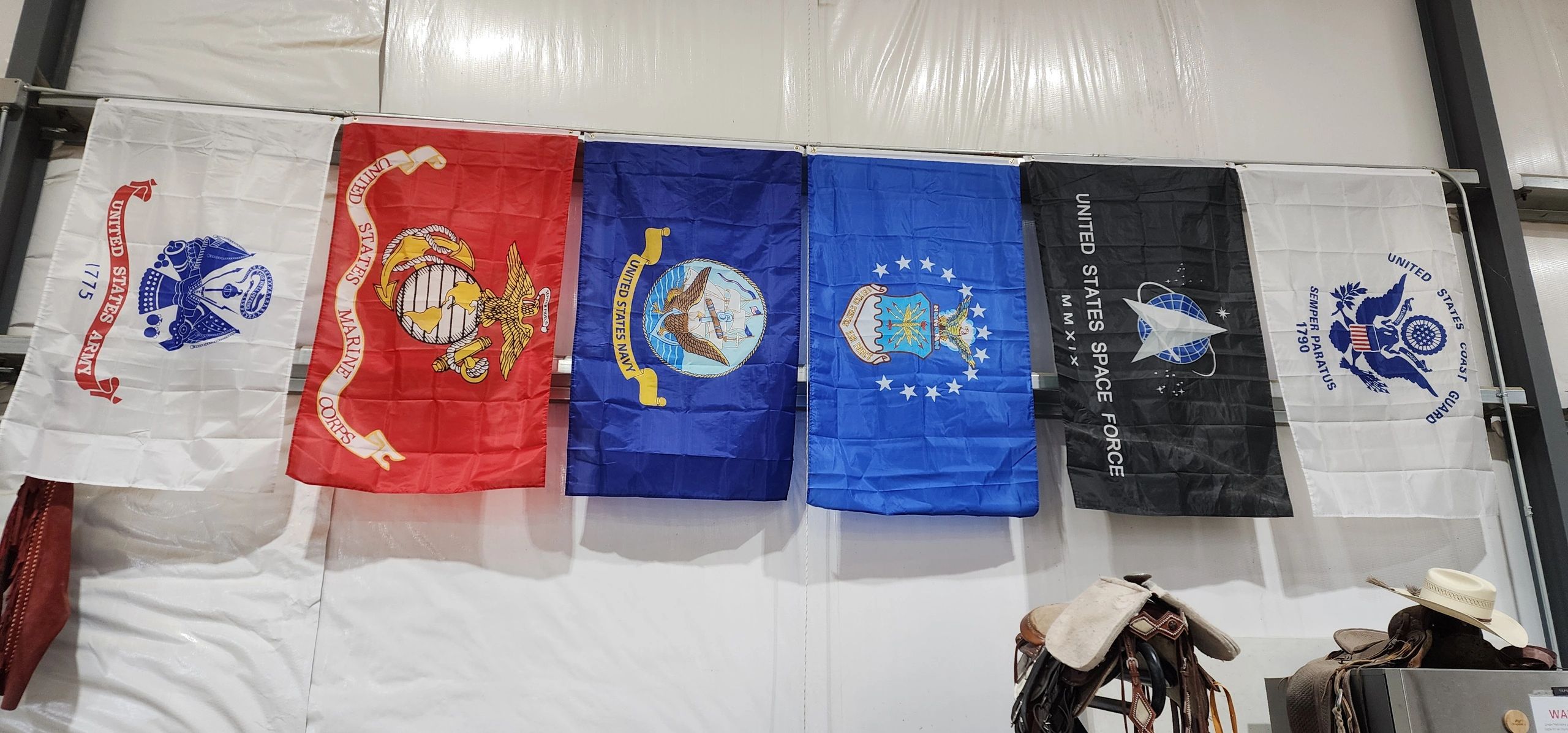Six U.S. military branch flags hanging indoors above saddles and a hat.