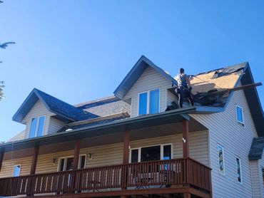 A worker repairs the roof of a beige house under clear blue sky.