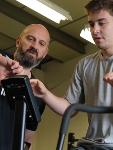 Trainer guiding a young man on exercise equipment in a gym.