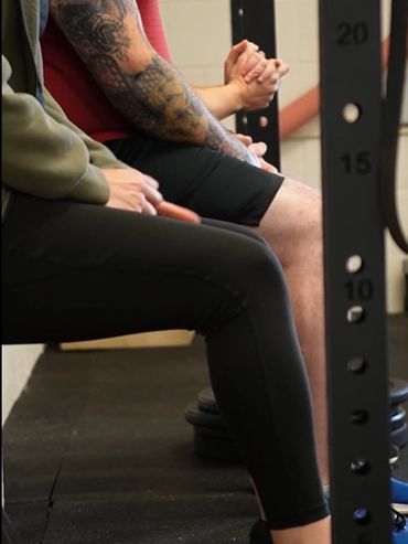 People sitting close together in a gym setting, showing tattooed arms and casual workout attire.