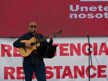 Man playing guitar and singing on stage with red and white resistance banner.