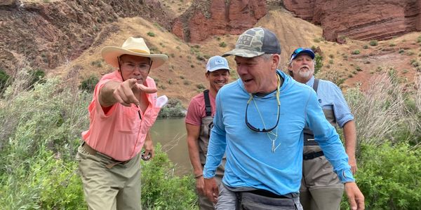 Four men in fishing gear posing playfully outdoors near a river and rocky hills.