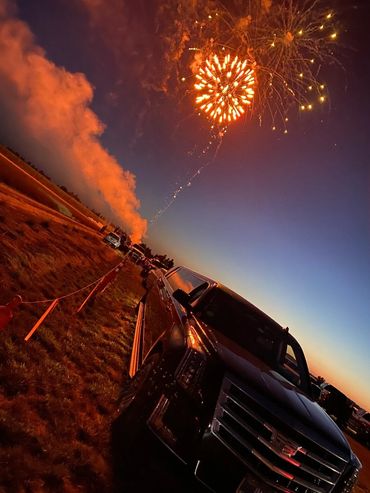 Fireworks light up the night sky above parked cars at sunset.