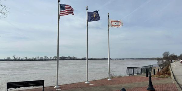 Three flags waving by a riverside promenade on a cloudy day.