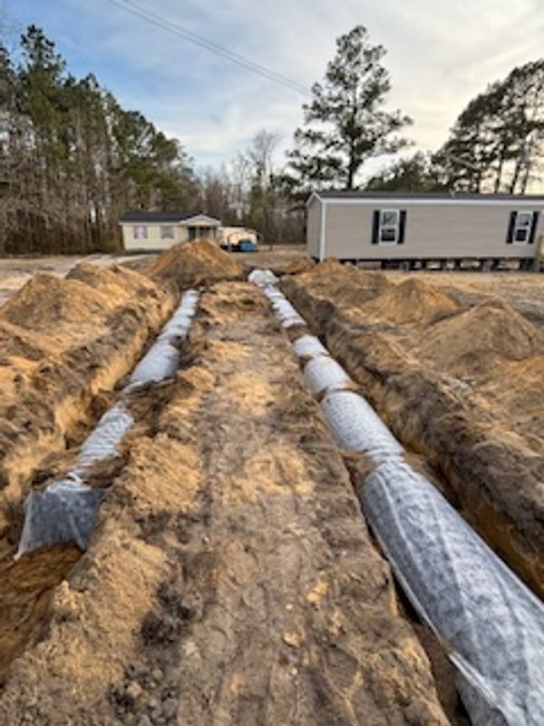 Construction site with trenches and wrapped pipes near houses and trees.